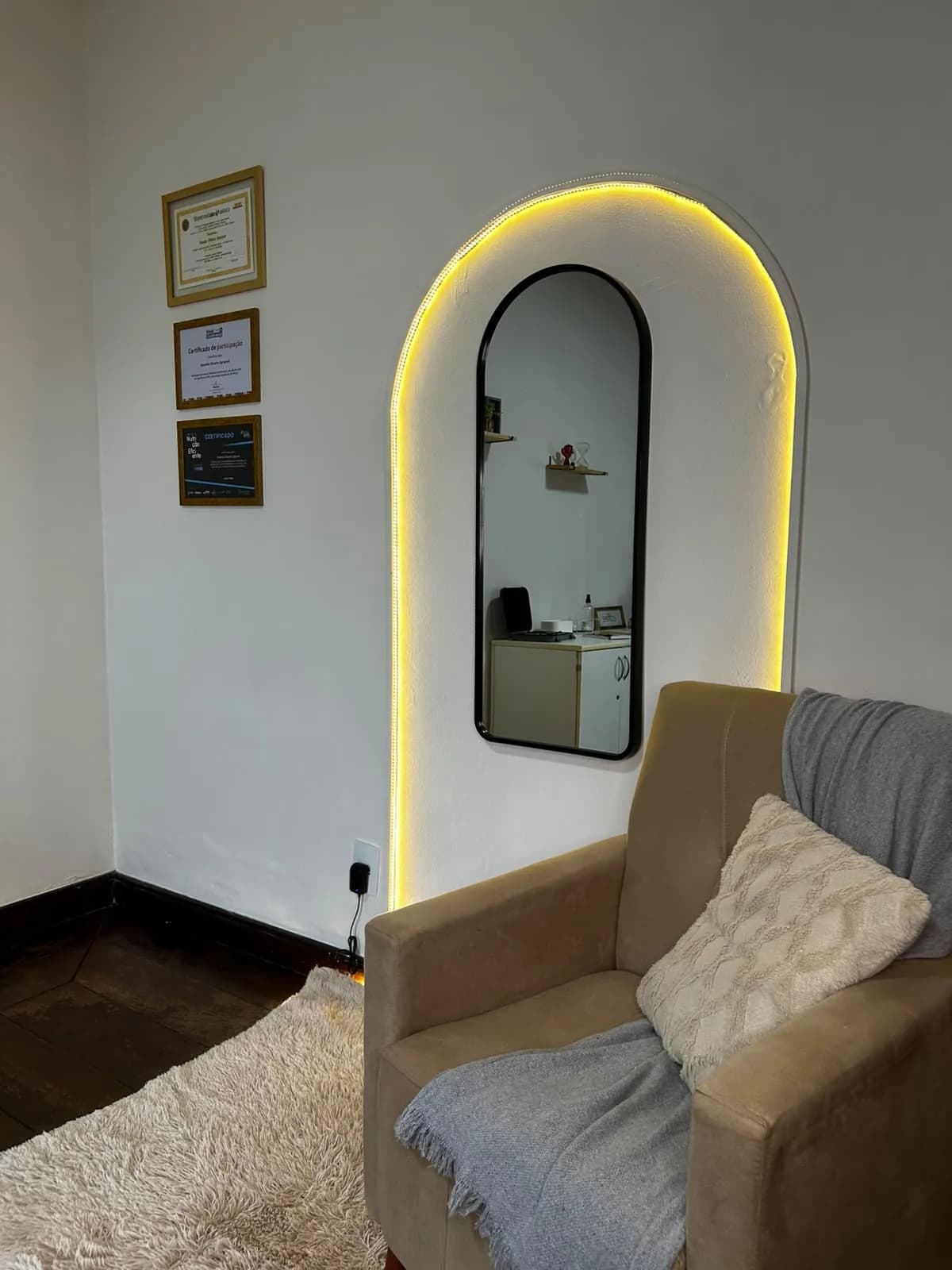 Cantinho de espera do consultório, com poltrona, espelho iluminado e certificados na parede
